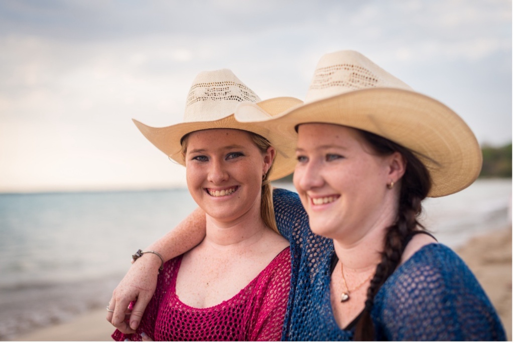 Two young women standing on a beach, both wearing pale coloured cowboys hats. One has her arm around the others' shoulder