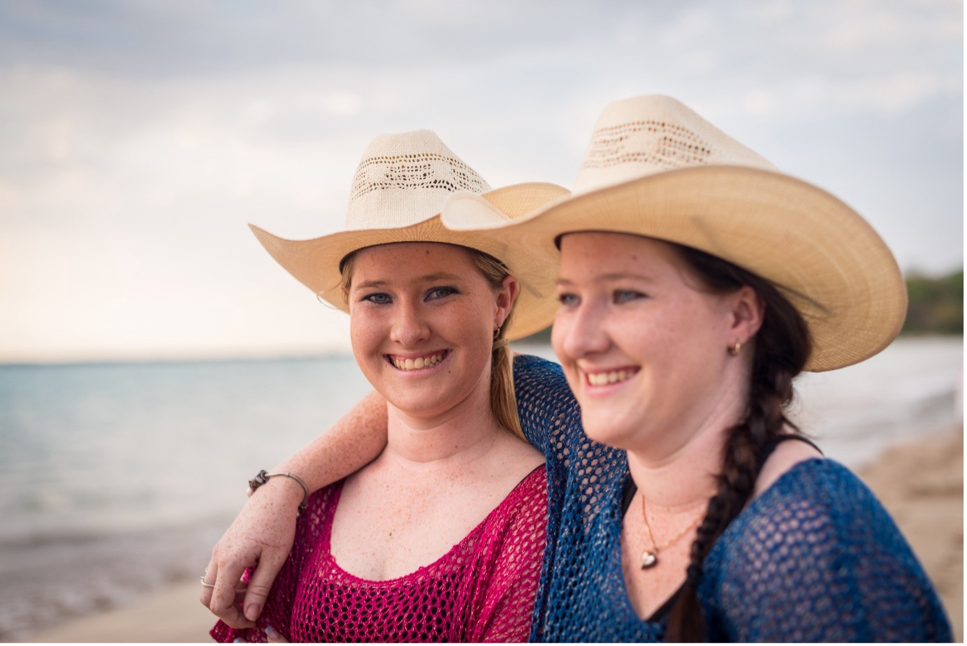 Two young women standing on a beach, both wearing pale coloured cowboys hats. One has her arm around the others' shoulder